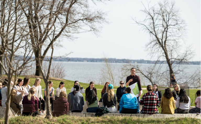 Omar Poler stands in a circle of students by the lake